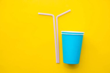 Sarı arka planda renkli plastik pipetler içen mavi kağıt bardaklar. Partiye hazırız. Üst Manzara. Minimalist stil. Kopyala, metin için boşluk.