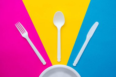 Plastik beyaz çatal, bıçak, kaşık ve plaka çok renkli arka planda. Mutfak aletleri. Üst Manzara. Minimalist stil. Kopyala, metin için boşluk.