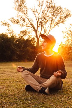 Meditasyon pozisyonunda genç yakışıklı bir adam ormanın ve günbatımının arka planında çimlerde oturur. Özgürlük rahatlama kavramı. Metin veya reklam için yer.