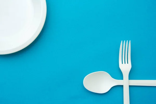 Plastic white crossed fork, spoon and plate on blue background. Cooking utensil. Cutlery sign. Top view. Minimalist Style. Copy, empty space for text.