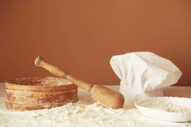 Masaya saçılmış beyaz buğday unu, ahşap merdane, kırsal elek ve kahverengi arka planda aşçı şapkası. Mutfakta lezzetli kurabiyeler pişir. Mutfakta rahat bir atmosfer var. Yemek yapmak için mutfak gereçleri.