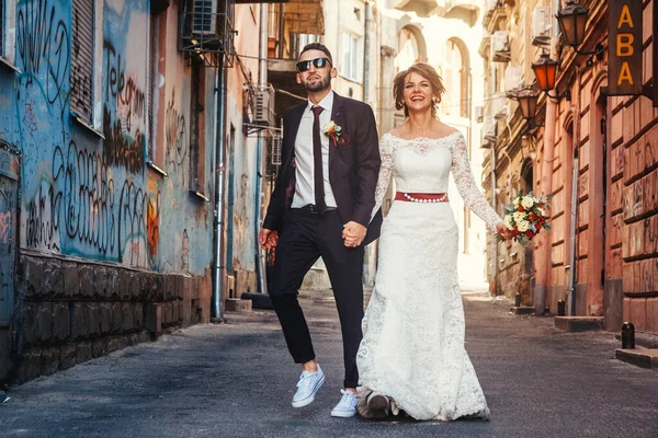 Young wedding couple celebrating marriage in city. Fun bride and groom smiling and walking in on modern city\'s street. Contemporary wedding.