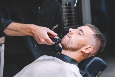 Genç erkek berber, müşterinin sakalını elektrikli makasla tıraş eder. Genç sakallı adam berber dükkanında kuaför tarafından tıraş ediliyor. Reklamcılık ve berber dükkanı konsepti.