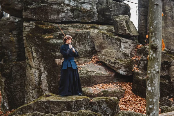 Young man in black kimono practicing martial arts with a sword on rocks and forest background. Place for text or advertising.