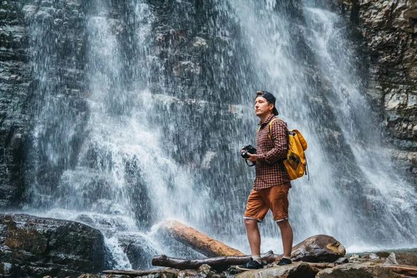 Traveler man with a yellow backpack standing on the background of a waterfall makes a photo landscape. Travel lifestyle concept. Copy, empty space for text