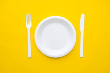 Plastik beyaz çatal, sarı arka planda bıçak ve plaka. Mutfak aletleri. Üst Manzara. Minimalist stil. Kopyala, metin için boşluk.
