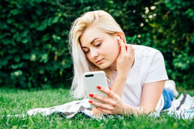 Kulaklıklı, akıllı telefonlu güzel sarışın kadın yeşil arka planda uzanan müziği dinliyor. Yaşam tarzı konsepti. Kopyala, metin için boşluk.