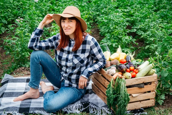Beautiful woman farmer sitting on a blanket near fresh organic vegetables in a wooden box against the background of a vegetable garden. Copy, empty space for text.