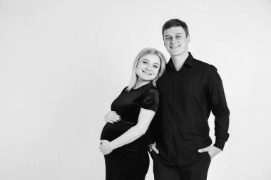 Hamile bir kadın ve kocası, beyaz arka planda siyah giysiler içinde karnına sarılıp sarılıyorlar. Siyah beyaz resim. Güzel evli bir çift. Kopyala, metin için boşluk.