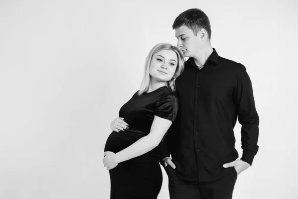 怀孕的女人和她的丈夫抱着你的肚子 穿着黑色衣服 背景是白色的 黑白照片 美丽的夫妇 空的文字空间 — 图库照片
