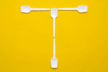Beyaz plastik kaşıklar sarı arka planda T harfi şeklinde. Üst Manzara. Kopyala, metin için boşluk.