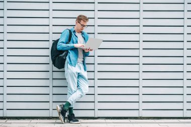 Gömlekli, kot pantolonlu ve gözlüklü, dizüstü bilgisayarını kullanarak tahta arka planda duran bir sırt çantası taşıyan yakışıklı bir genç. Serbest çalışma konsepti. Kopyala, metin için boşluk.