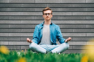 Gömlekli, kot pantolonlu ve gözlüklü genç adam gri bir duvarın arka planında yeşil çimlerde meditasyon pozunda oturuyor. Kopyala, metin için boşluk.