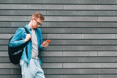 Gömlekli, kot pantolonlu ve gözlüklü genç bir adam sırt çantası taşıyor ve arka planda gri bir duvarın üzerinde dururken akıllı telefona bakıyor. Kopyala, metin için boşluk.