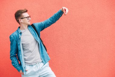 Gömlekli, kot pantolonlu ve gözlüklü genç bir adam sırt çantası taşıyor ve pembe bir duvarın arka planında cep telefonuyla selfie çekiyor. Kopyala, metin için boşluk.