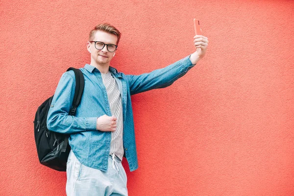 Portrait young man in shirt, jeans and glasses carrying a backpack and looking at smartphone while standing on a pink background. Copy, empty space for text.