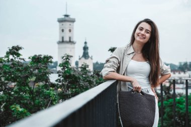 Beyaz elbiseli, elinde örgü çantalı, şehrin caddesinde poz veren güzel esmer bir kadın. Kopyala, metin için boşluk.