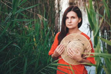 Sazlık ve gölün arka planında kırmızı giysiler giymiş hasır şapkalı güzel bir kadının portresi. Kopyala, metin için boşluk.