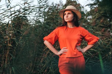 Sazlıkların arka planında kırmızı giysiler giymiş hasır şapkalı güzel bir kadın. Kopyala, metin için boşluk.