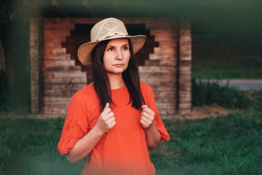 Hasır şapkalı güzel bir kadın ahşap bir evin arka planında duruyor. Kopyala, metin için boşluk.