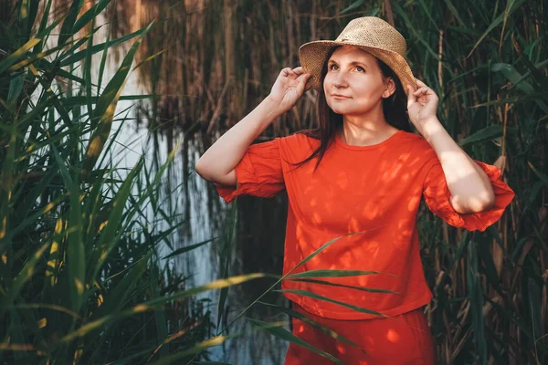 Portrait of a beautiful woman in a straw hat dressed in red clothes on a background of reeds and lake. Copy, empty space for text.