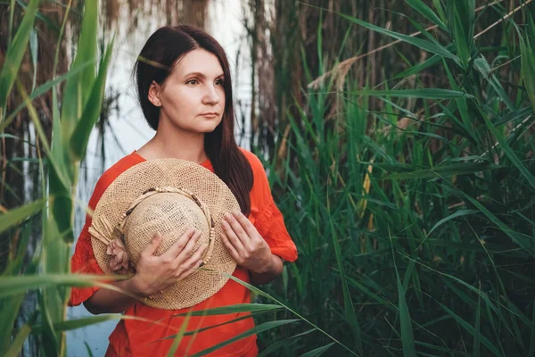 Portrait of a beautiful woman in a straw hat dressed in red clothes on a background of reeds and lake. Copy, empty space for text.