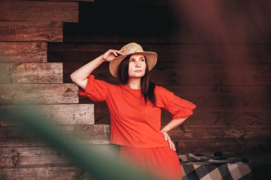 Hasır şapkalı güzel bir kadın ahşap bir evin arka planında duruyor. Kopyala, metin için boşluk.