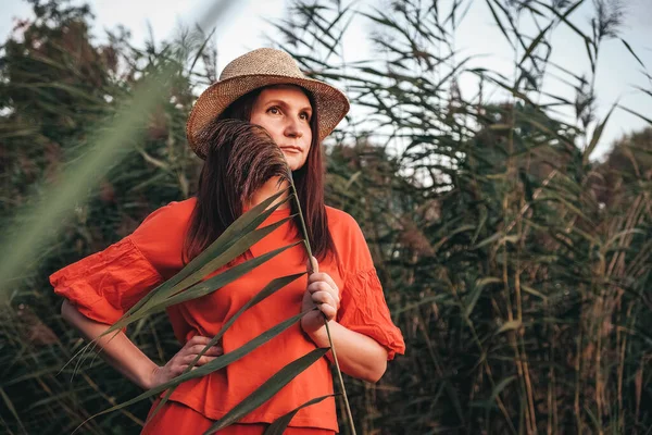 Portrait of a beautiful woman in a straw hat dressed in red clothes on a background of reeds and lake. Copy, empty space for text.