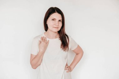 Güzel esmer kadın ellerini yanağında tutuyor, günlük beyaz tişört giyiyor, çok az makyaj yapıyor, beyaz arka planda içeride poz veriyor. Kopyala, metin için boşluk.