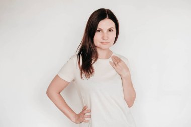 Güzel esmer kadın ellerini yanağında tutuyor, günlük beyaz tişört giyiyor, çok az makyaj yapıyor, beyaz arka planda içeride poz veriyor. Kopyala, metin için boşluk.