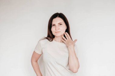 Güzel esmer kadın ellerini yanağında tutuyor, günlük beyaz tişört giyiyor, çok az makyaj yapıyor, beyaz arka planda içeride poz veriyor. Kopyala, metin için boşluk.