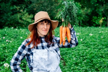 Güzel bir kadın çiftçinin portresi hasır bir şapka içinde bir demet havuç tutuyor ve sebze bahçesindeki bitkilerle çevrili..