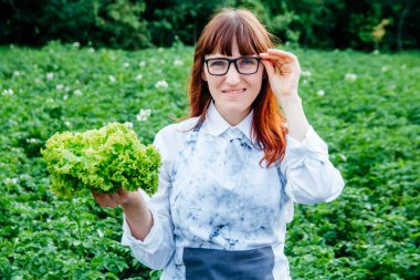 Yeşil lahana yaprakları tutan güzel bir kadın çiftçi, kameraya gülümsüyor ve sebze bahçesinde bir sürü bitkiyle çevrili..
