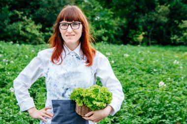 Hasır sepetinde yeşil lahana yaprakları tutan, kameraya gülümseyen ve sebze bahçesinde bir sürü bitkiyle çevrili güzel bir kadın çiftçi..