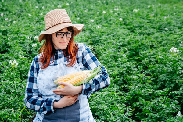 Portrait of a beautiful female farmer holding a corns cobs in a straw hat and surrounded by plenty of plants in her vegetable garden.