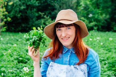 Kadın çiftçi portresi elinde bir demet maydanozla hasır şapka tutuyor ve etrafını sebze bahçesindeki bitkilerle çeviriyor..