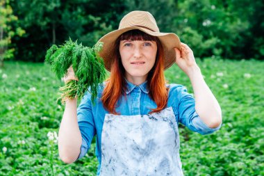 Kadın çiftçi portresi elinde bir demet maydanozla hasır şapka tutuyor ve etrafını sebze bahçesindeki bitkilerle çeviriyor..