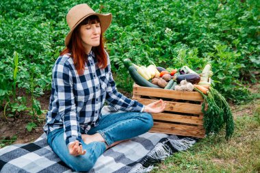 Güzel bir kadın çiftçi, sebze bahçesinin arka planındaki tahta bir kutuda taze organik sebzelerin yanında battaniyenin üzerinde otururken meditasyon yapıyor..