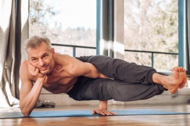 Çıplak gövdesi olan yaşlı bir adam stüdyoda pencerenin önünde yoga yapıyor. Kopyala, metin için boşluk.