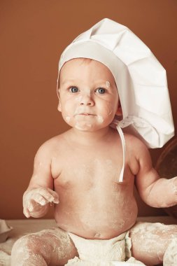 Şef şapkası takmış küçük fırıncı çocuk kahverengi arka planda un ile ahşap bir raptiye, yuvarlak bir süzgeç ve yumurtalarla oynuyor. Kopyala, metin için boşluk.