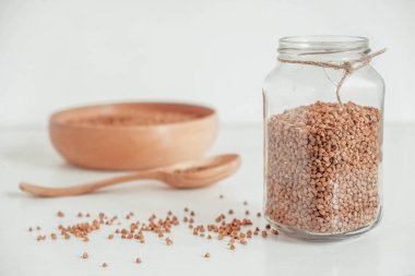 Cam kavanozda karabuğday taneleri, beyaz arka planda ahşap kase ve kaşık. Kopyala, metin için boşluk.