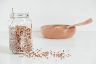 Cam kavanozda karabuğday taneleri, beyaz arka planda ahşap kase ve kaşık. Kopyala, metin için boşluk.