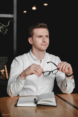 Masada elinde kalemle oturan beyaz gömlek ve gözlük takan genç adam, ofisteki karanlık geçmişiyle ilgili notlar alıyor. Kopyala, metin için boşluk.