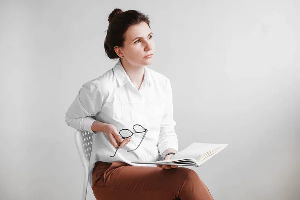 Красива жінка в окулярах і одягнена в білу сорочку сидить на стільці з книгою в руках на білому тлі. Копіювати, порожній простір — стокове фото