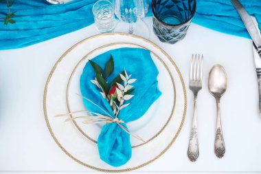 Mavi kumaşla kaplı beyaz ziyafet masası, tabaklar, çatal bıçak takımı, bardaklar, mumlar ve çiçek aranjmanları ile servis ediliyor. Üst Manzara. Kopyala, metin için boşluk .