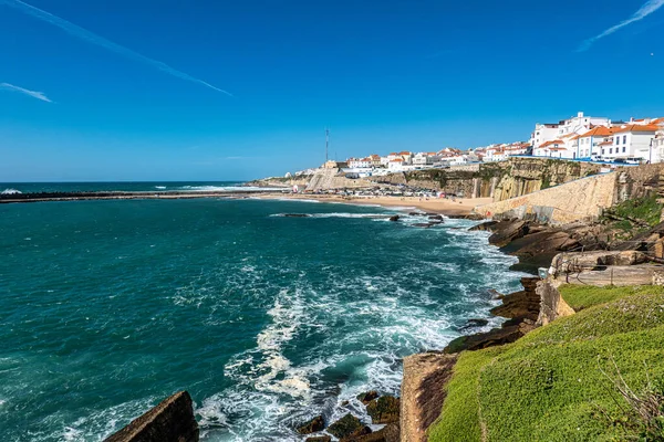 Portekiz 'in Ericeira kentindeki Praia do Sul manzarası altın kum, turkuaz sular ve canlı bir gökyüzü altında kıyı binaları.