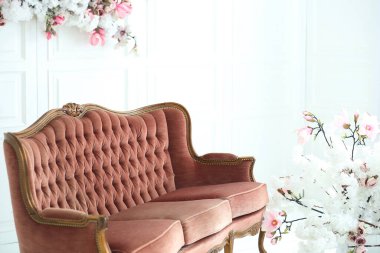 Güneşli bir günde odada klasik tarzda rahat bir kanepe, beyaz bir duvarda suni çiçeklerden oluşan çiçek dekoru. Tasarım ve iç mekan konsepti. Boşluğu kopyala