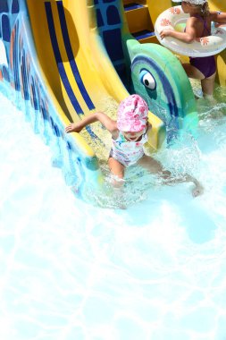 Pembe şapkalı ve mayolu neşeli beyaz bir kız su kaydırağından havuza kayıyor. Denizde çocukların eğlencesi. Yaz ve deniz. Çocuk konseptiyle tatil.