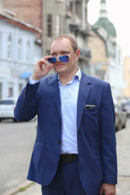 Mavi takım elbiseli genç, kendinden emin beyaz adamın portresi elinde güneş gözlüğü tutarken ileriye bakıyor. Arka plan şehrin hareketli iş merkezidir. Avrupa. İş, iş.