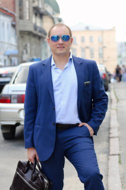 Mavi takım elbiseli, elinde evrak çantası tutan güneş gözlüklü, kendinden emin beyaz bir gencin portresi. Arka plan şehrin hareketli iş merkezidir. Avrupa İş, İş.